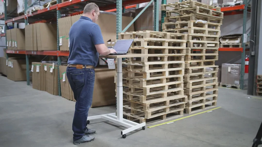 The photo shows a person working on a laptop at a height-adjustable desk, in a large, orderly warehouse filled with cartons and wooden pallets. Industrial racking is visible in the background. - Photo: TheStandingDesk