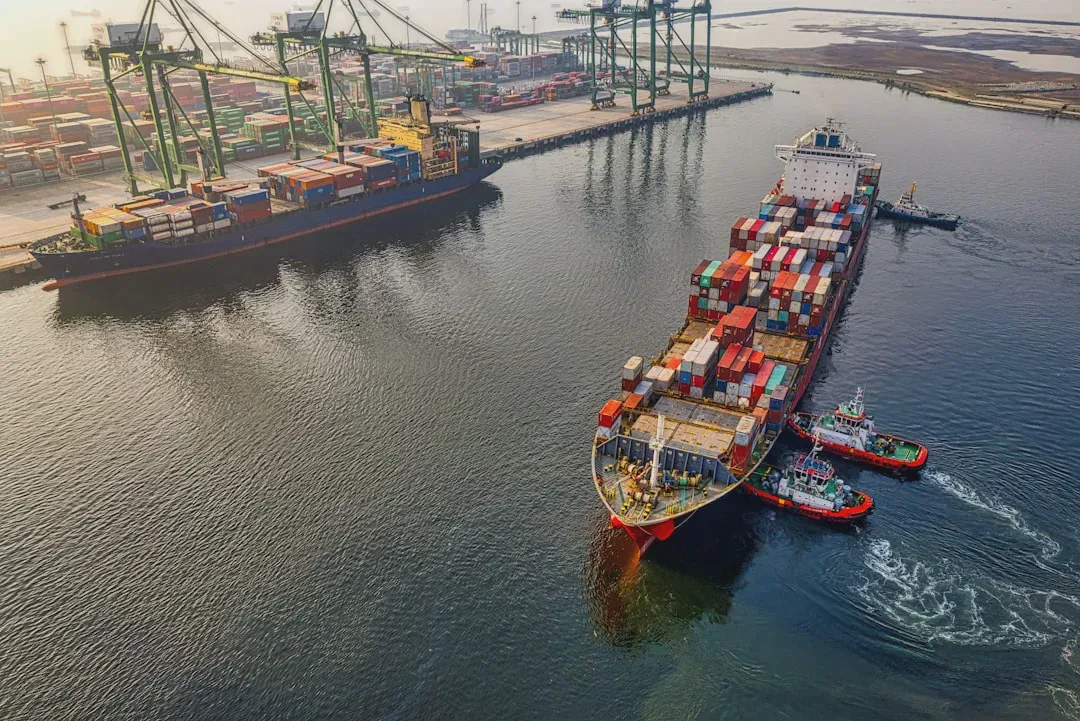The photo shows a bustling port with large cargo ships loaded with containers, surrounded by smaller tugboats. The colorful containers and activity around the vessels give the photo a dynamic character. - Photo: william william