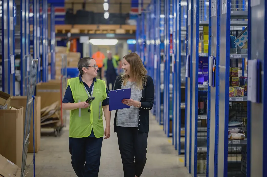 The photo shows two people in a well-lit, orderly warehouse. One is wearing a hi-vis vest and holding a scanner, the other is holding a notepad. Blue racking filled with products indicates an active logistics operation. - Photo: Centre for Ageing Better