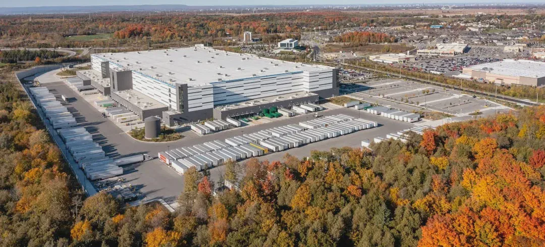 The photo shows a large, modern warehouse complex surrounded by autumn greenery. Amazon-branded trucks are visible in the scene, suggesting a fulfillment center or logistics hub. - Photo: Point3D Commercial Imaging Ltd.